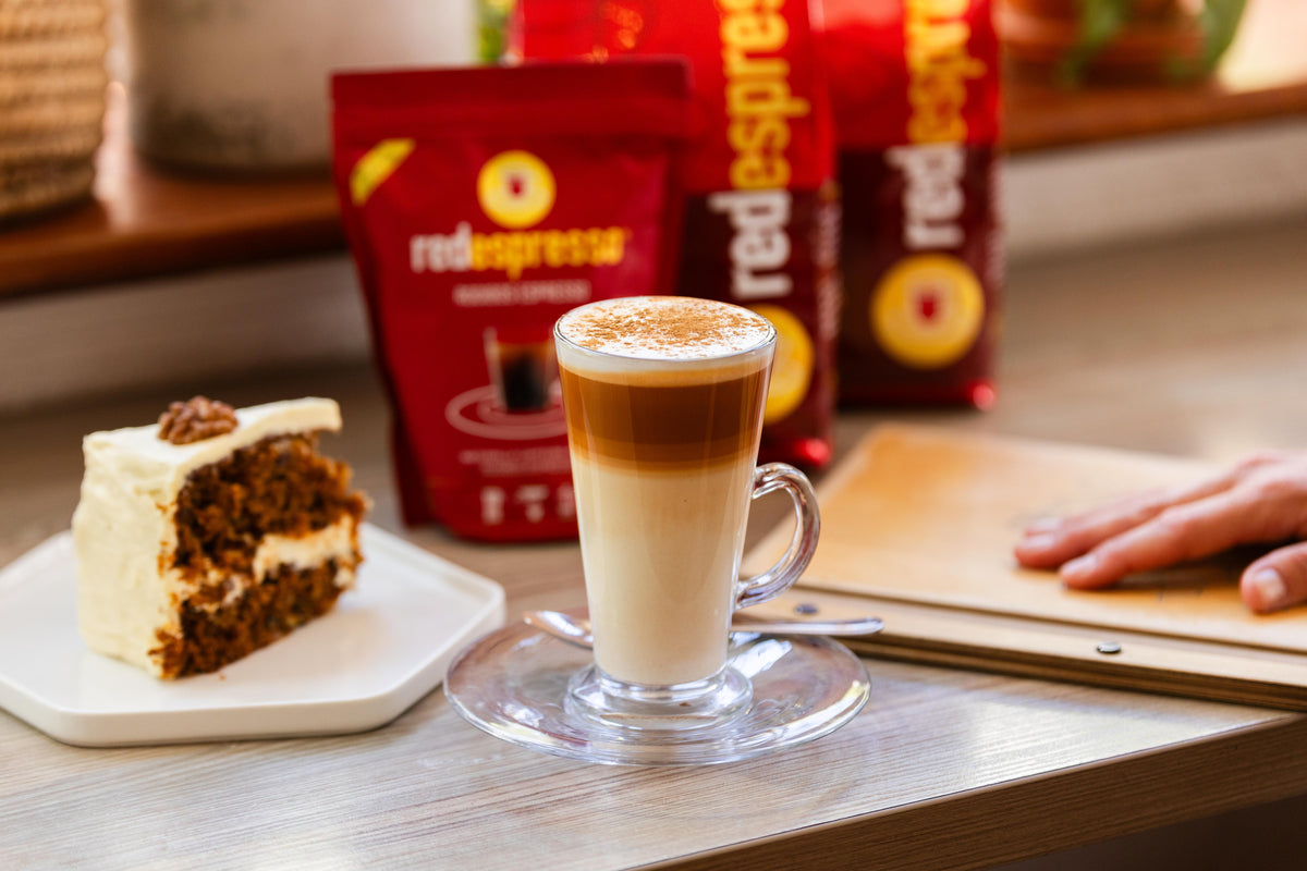 Rooibos Red Latte® mit Kuchenstück sowie Red Espresso Packungen auf Tisch in einem Café